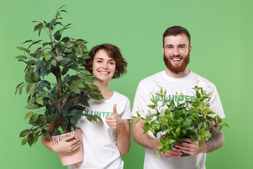 Smiling two young friends couple in white volunteer t-shirt isolated on green background. Voluntary free work assistance help charity grace teamwork concept. Hold green flowerpots, showing thumb up.