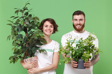 Smiling two young friends couple in white volunteer t-shirt isolated on pastel green background in studio. Voluntary free work assistance help charity grace teamwork concept. Hold green flowerpots.