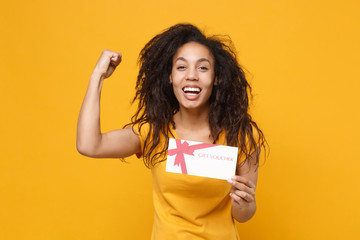 Fototapeta na wymiar Laughing young african american woman girl in casual t-shirt posing isolated on yellow orange wall background. People lifestyle concept. Mock up copy space. Hold gift certificate doing winner gesture.