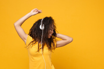 Fototapeta premium Cheerful young african american woman girl in casual t-shirt posing isolated on yellow orange background in studio. People lifestyle concept. Mock up copy space. Listen music with headphones, dancing.