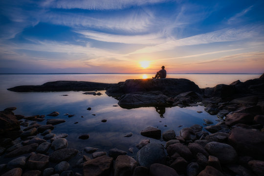 Man sitting on a cliff by a lake during sunset