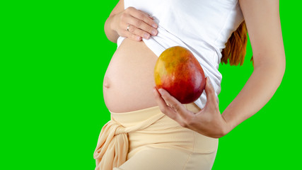 A happy pregnant young woman holding a mango next to her belly showing the size of the baby inside her belly. Photos of fetal growth at 19 weeks pregnancy. Healthy pregnancy diet with vitamins