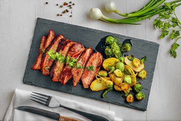 Gourmet meal made of hanger steak, brussels sprouts, potatoes and onion on a stone board. Tasty medium rare meat steak with vegetables on a white wooden table. Top view above.