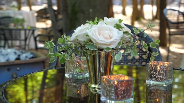 Romantic garden table decoration. Vase with white roses and candles in square shaped glass. Trees reflecting on the table surface. Parallax shot.