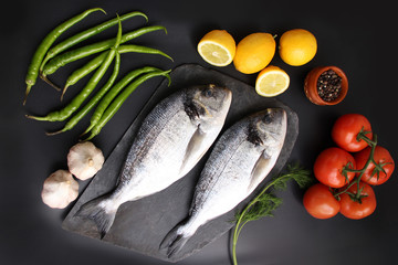 lemons, tomatoes, hot peppers and two carcasses fresh raw atlantic saltwater fish Sparus aurata with shiny silver scales lie on a black background, cooking concept