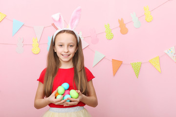 Obraz na płótnie Canvas Portrait of a little cute smiling girl with bunny ears and easter eggs in hands on a colored background. Easter background with place to insert text.