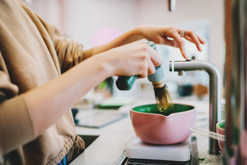Process of preparing traditional green matcha tea. Barista adding water in a bowl for the matcha tea. Woman's hand holding modern electric whisk for whipping the matcha powder in coffee shop.