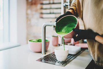 Matcha tea made of natural ingredients by professional barista in asian cafe. Female's hands holding a bowl with a freshly made matcha pouring to the white cup. Tea ceremony in an asian style.