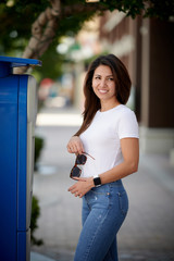 Smiling woman in white T-shirt and blue jeans standing on the street next to parking holding sunglasses