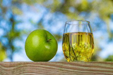 Apple juice. Organic green apple. Fruit drink in a glass. Wooden table. Beautiful background with blue sky and green tree leaves.