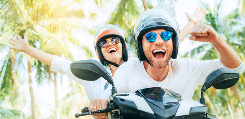 Happy smiling couple travelers riding motorbike scooter in safety helmets during tropical vacation under palm trees on Ko Samui island , Thailand