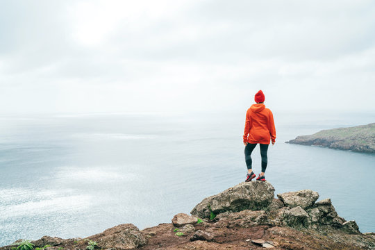 Trail runner female dressed orange sporty hoodie and red cap resting on the cliff and enjoying Atlantic ocean view on Ponta de Sao Lourenço peninsula -the easternmost point of Madeira island, Portugal