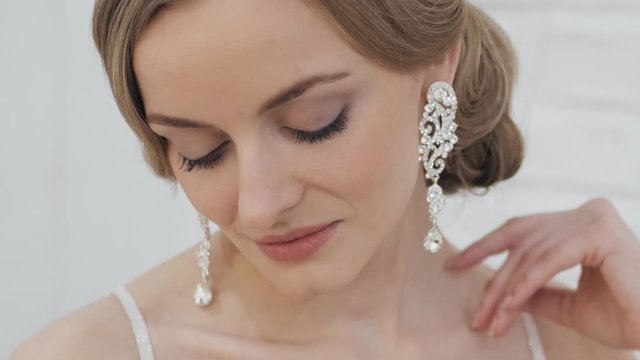 Big close up for social networks stories of young blonde bride with wedding hairstyle in tiara earrings wedding gown with bare shoulders posing in trendy wedding salon turning head teasing and