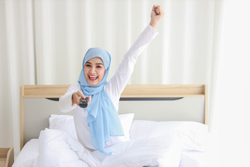 TV and happiness concept. Beautiful asian muslim woman in white sleepwear sitting on bed, holding television remote and raise hand, shout loudly and celebrate something with glad happy smiling face.