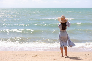 Fototapeta na wymiar Rear view happiness traveller young asian woman in blue dress and hat standing on sandy beach. Cute girl enjoy her tropical sea on her holiday vacation during summer time and sunshine day.