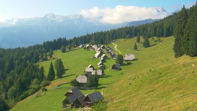 DRONE: Scenic view of a small village of cottages with a view of the stunning Julian Alps. Picturesque forest and untouched forest surrounds a row of wooden huts near a hiking trail in Slovenia.