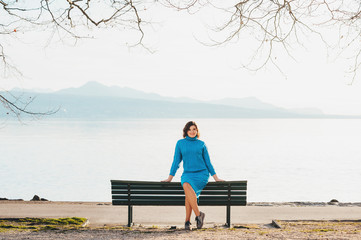 Fototapeta premium Outdoor portrait of young woman resting on the bench by the lake in early spring, wearing blue knitted pullover dress