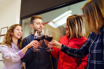 Group of caucasian friends or family standing by the table at winery or restaurant holding a glasses of red wine toasting celebrating young man and three women wearing shirt smiling