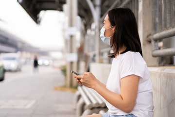 Asian young woman waiting for the bus at bus stop in the crowded and busy city and touching on smartphone for searching a map. City where is fully with air pollution and crowd. Environment problem.