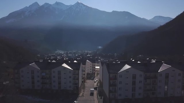 Aerial view of the small town situated in the valley surrounded by high mountains covered by trees. Clip. Flying above the town near forested hills on blue sky background.