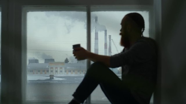 Portrait of a red-haired man with a beard sitting by the window against the background of pipes emitting smoke.