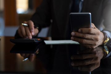 Businessman in suit,hand using mobile smart phone and calculating business budget and incomes of company at modern office.