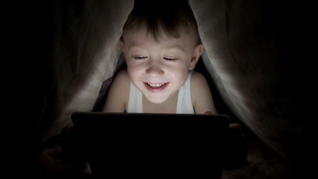 little boy plays on tablet while lying under blanket. watching cartoons hiding