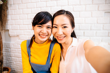 Face of smiling happy Asian teenage daughter and Asian middle-aged mother looking at mobile phone in indoor living room at home. It could be video calling or taking a selfie photo together.