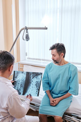 Obraz premium Sad mature patient with cancer sitting on bed and listening to doctors recommendations while doctor viewing x-ray image