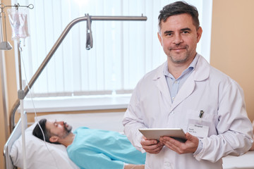 Obraz premium Portrait of content handsome middle-aged doctor with stubble using tablet, ill patient sleeping in background