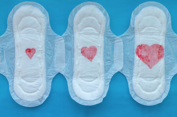 Women's pads with red hearts of different sizes on a blue background. The concept of the menstrual cycle.