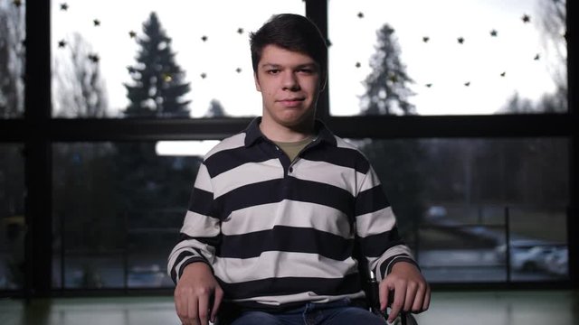 Close-up portrait of confident teenage boy with impaired mobility sitting in wheelchair in sport hall. Smiling teenager with cerebral palsy posing for camera indoors