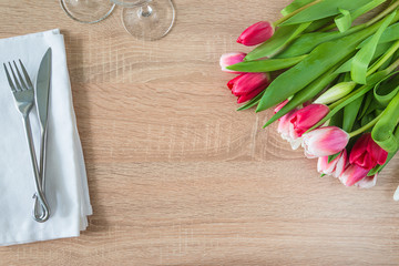 Festive table setting with bunch of purple tulips, forged fork and knife and set of wineglasses on white napkin and wooden background. Space for text, top view