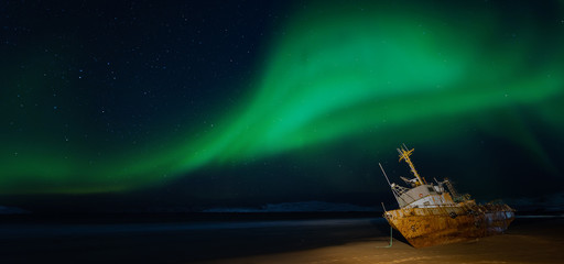 Polar lights in the starry sky. A fishing boat lying on its side, washed up by a storm on the Bank of the Barents sea. Teriberka, Russia