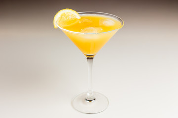An orange cocktail with ice in a martini glass garnished with lemon