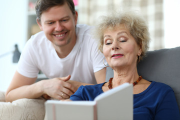 Elderly lady sits sofa watches album with her son. Productive and interesting time with an elderly mom. Adult son takes into account need for parents to communicate. Son visits mother in nursing home