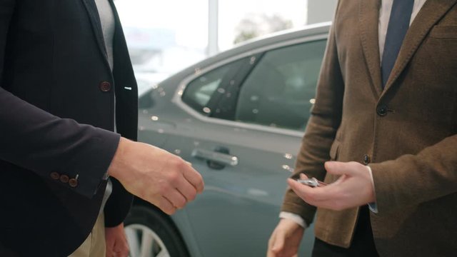 Slow Motion Close-up Of Anonymous Men Shaking Hands In Car Showroom Taking Key Fob Walking To New Automobile. Agreement, People And Business Concept.