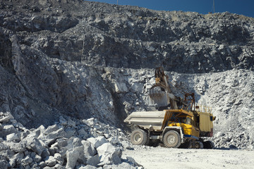 Mining excavator and heavy mining truck in a quarry for the extraction of limestone on the background of rocky terrain in sunny weather, close-up. - obrazy, fototapety, plakaty