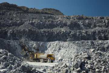 Mining excavator and heavy mining truck in a quarry for the extraction of limestone on the background of rocky terrain and blue sky in sunny weather.