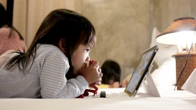 Adorable little girl lying on bed with sucking her thumb and looking on a digital tablet in  bedroom. 