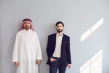 Arab and european businessmen stand smiling at the gray background.