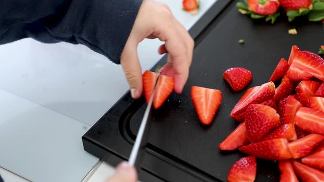 Close up of girl's hand removing strawberry leaves before slicing them in half with a knife. Pile of strawberry on black kitchen countertop