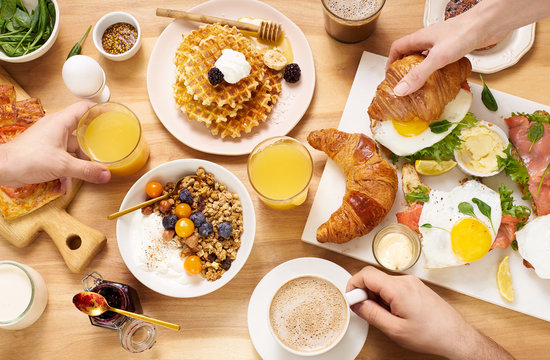 Top view image of brunch menu on wooden table with hands holding food and drinks. Healthy sunday breakfast with croissants, waffles, granola and sandwiches.