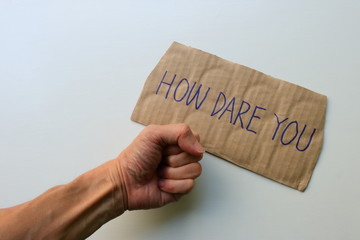 Closeup and focus on cardboard sign read HOW DARE YOU, next to clenched fist on white wooden background, climate change concept