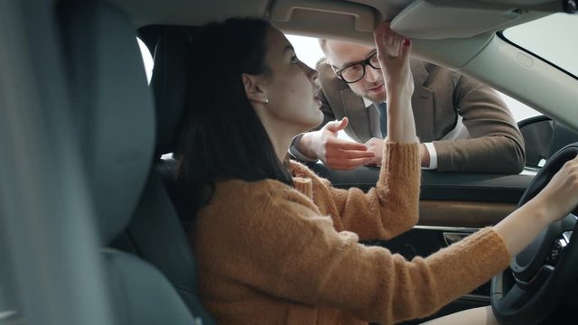 Pretty lady is talking to sales representative in motor showroom sitting inside brand new car holding steering wheel. Communication and deals concept.
