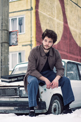 A bearded man in a coat sits on a car.