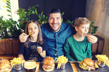 Modern dad with dental braces having fun with his kids eating hamburgers with french fries at trendy fast food restaurant - 328028659