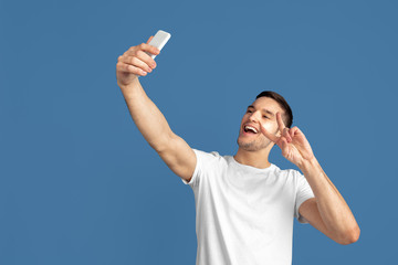 Taking selfie. Caucasian young man's portrait isolated on blue studio background. Beautiful male model in casual style, pastel colors. Concept of human emotions, facial expression, sales, ad.