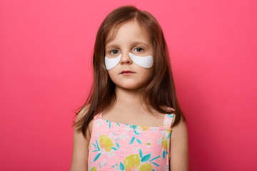 Portrait of emotional upset female child posing isolated over rose background, has white patches under eyes, standing with serious facial expression, looks directly at camera, wears t shirt or dress.