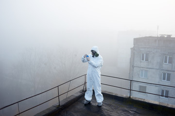 Human in protective suit holding a flask, is in the center of the snapshot in foggy morning, making research on air pollution study, working on roof of high building